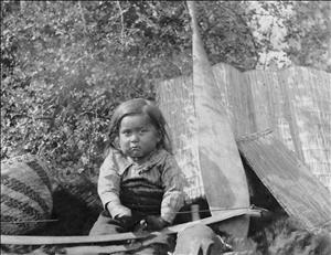 An indigenous child sits on the ground holding a long bow used for shooting arrows in his lap. Reed mats lean against shrubs behind him and a woven basket is off to one side. He is scowling or pouting. 